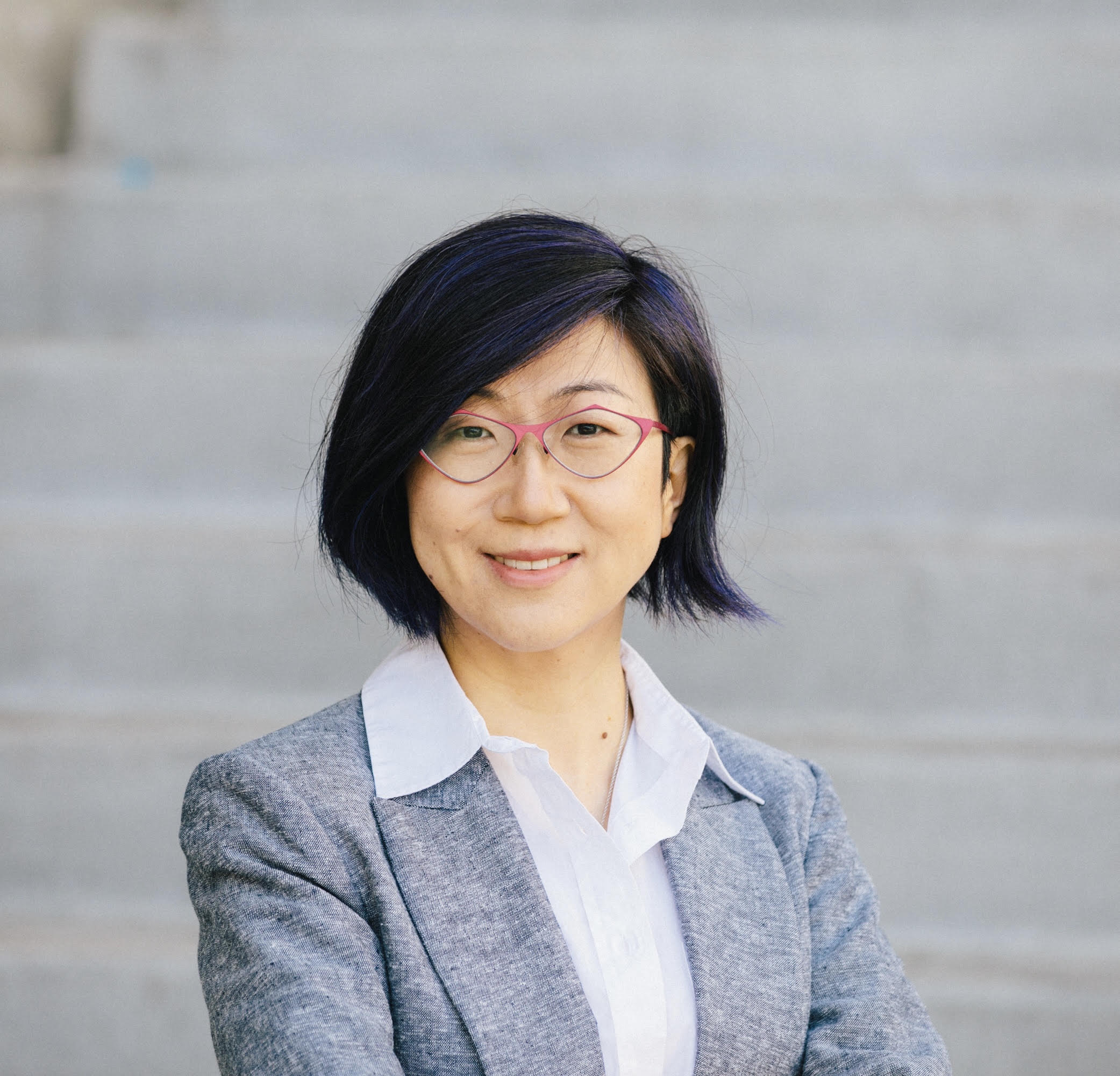 A professional headshot of a smiling Asian woman with chin-length dark brown hair and magenta glasses wearing a white collared blouse and blue-gray blazer against softly blurred stone steps.
