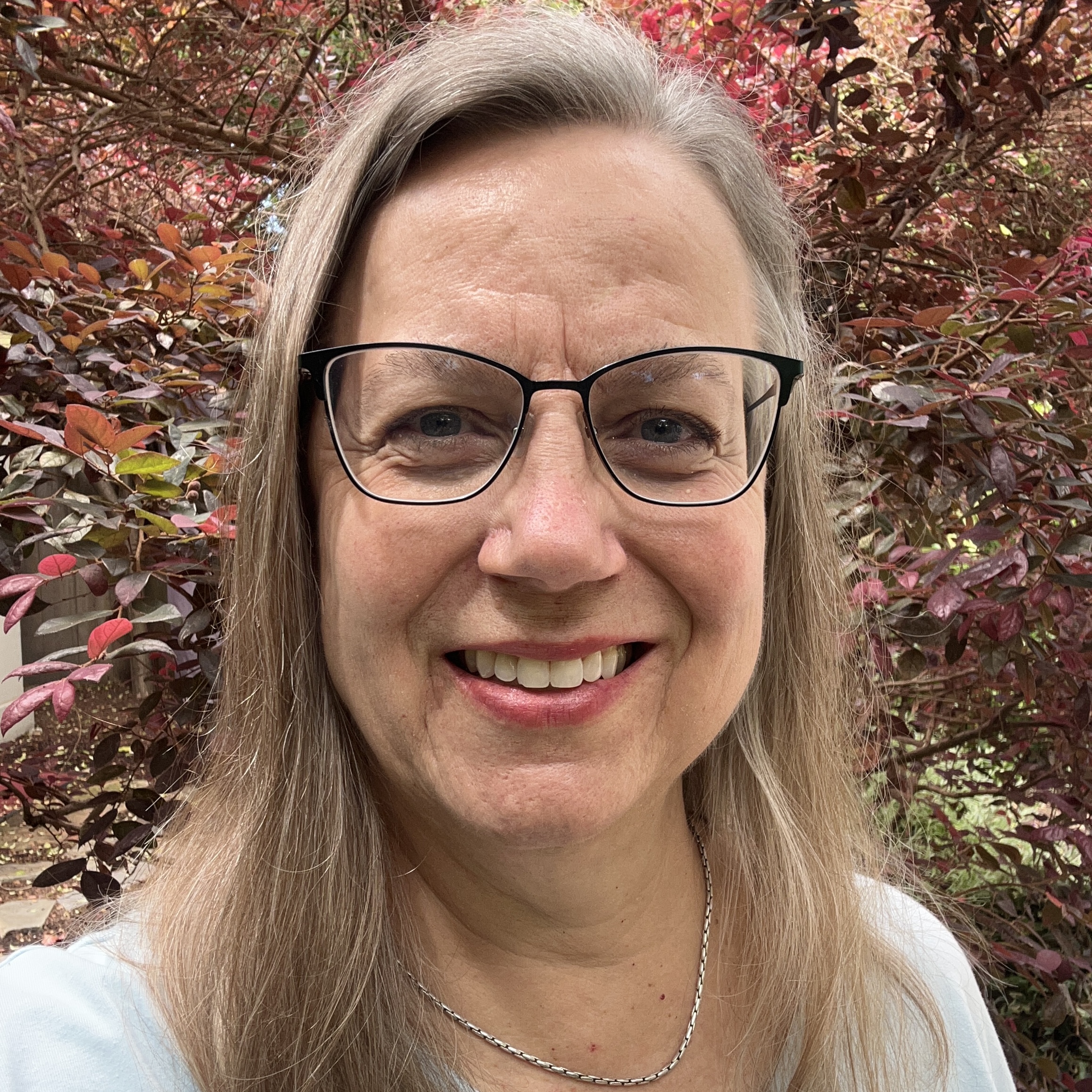 A smiling gray-haired woman with glasses poses in front of foliage.