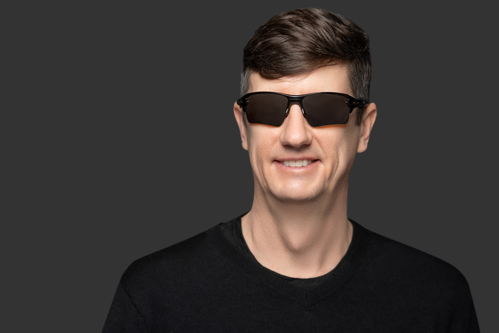 A smiling man with short brown hair wearing dark sunglasses and a black V-neck sweater.